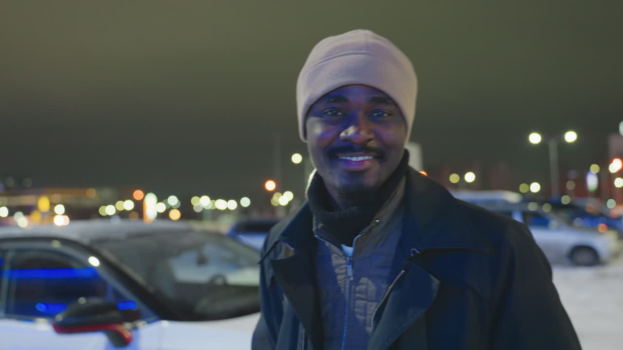 Young man dressed in winter coat and hat turns with warm smile on face while standing outside during cold night, illuminated by glowing city lights and snowy background