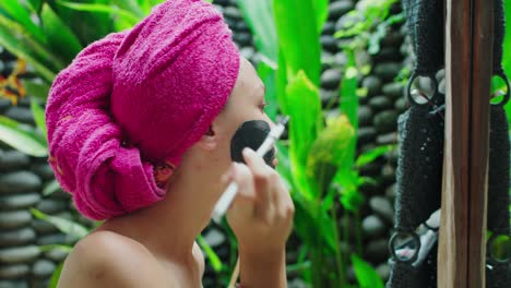 young woman with towel on head applying clay mask on her face after shower in tropical bali style bathroom