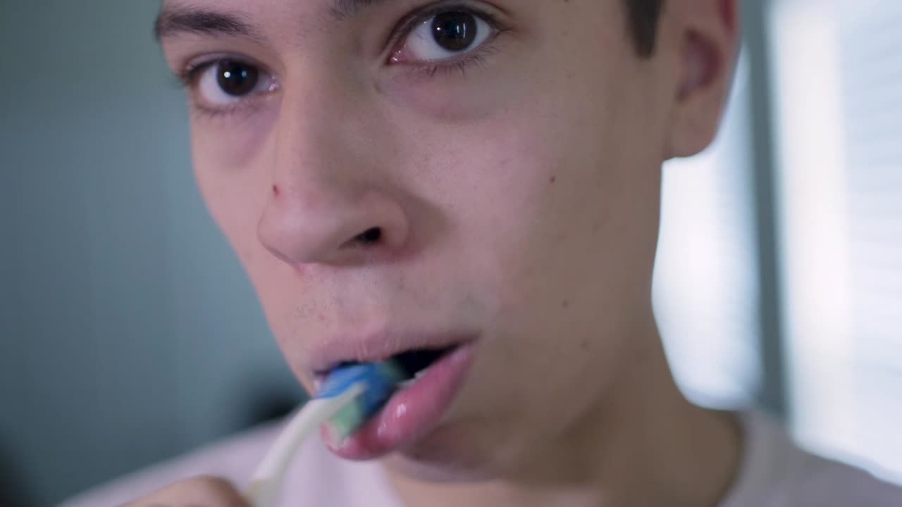 Young Caucasian Man Brushes Teeth Looking Straight at Camera Close Up