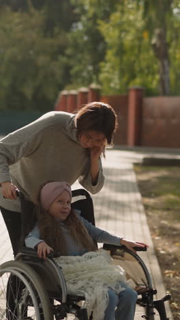 happy mom leans to kiss face of little daughter in wheelchair on stone pavement. woman and girl with injury enjoy walk in city park on sunny day