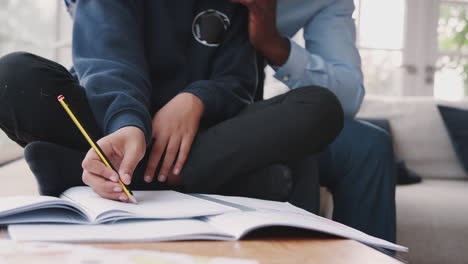 Close-up,-low-section-of-mixed-race-pre-teen-boy-sitting-cross-legged-doing-homework-with-his-dad-sitting-behind-helping-him