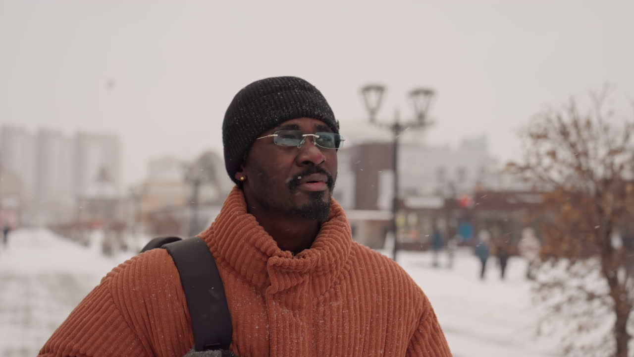 Uomo nero con maglione arancione che guarda attraverso una piazza innevata, viaggiatore indipendente che esplora il percorso, spallina dello zaino visibile, lampioni e pedoni sfocati, neve leggera che cade, ampia vista urbana, speranzoso