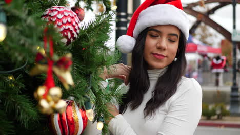 una mujer alegre con un sombrero festivo de santa claus decorando un árbol de navidad con adornos, cascabeles y luces