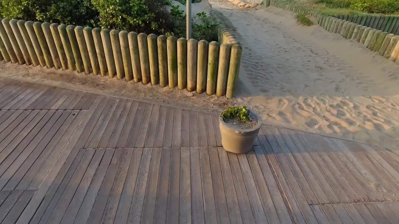 Panorering op mod et skilt på en træpæl ved siden af en sti med havet og horisonten i baggrunden, omgivet af grønne planter og et træhegn
