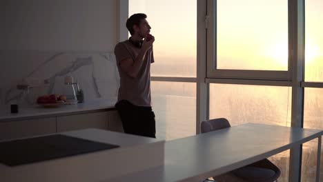 tall, young single man with headphones on neck relaxing by the window at studio kitchen, watching sunset and taking apple in his