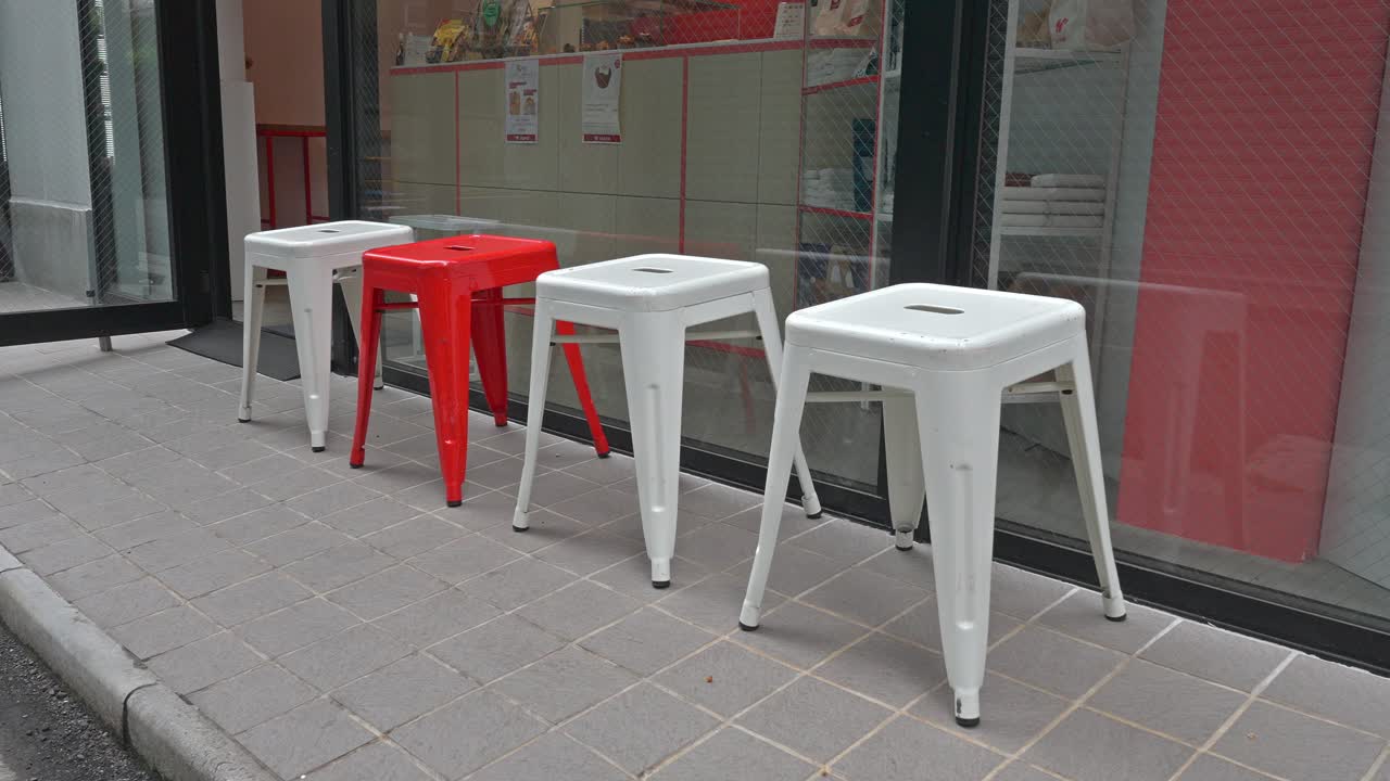 A visually striking arrangement of modern metal stools, prominently featuring one red stool among several white ones, positioned outside a clean, contemporary shopfront.