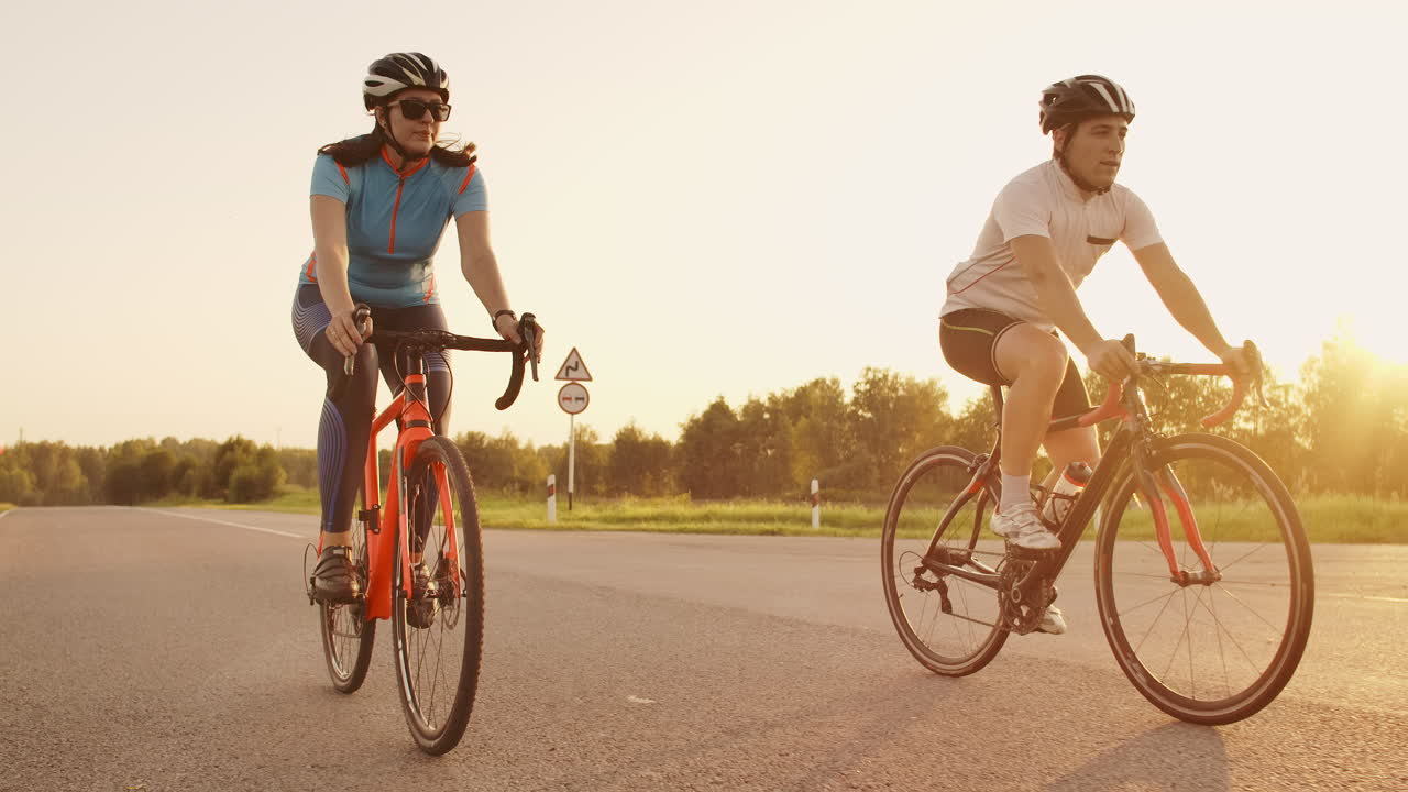 Two cyclists a man and a woman ride on the highway on road bikes wearing helmets and sportswear at sunset in slow motion