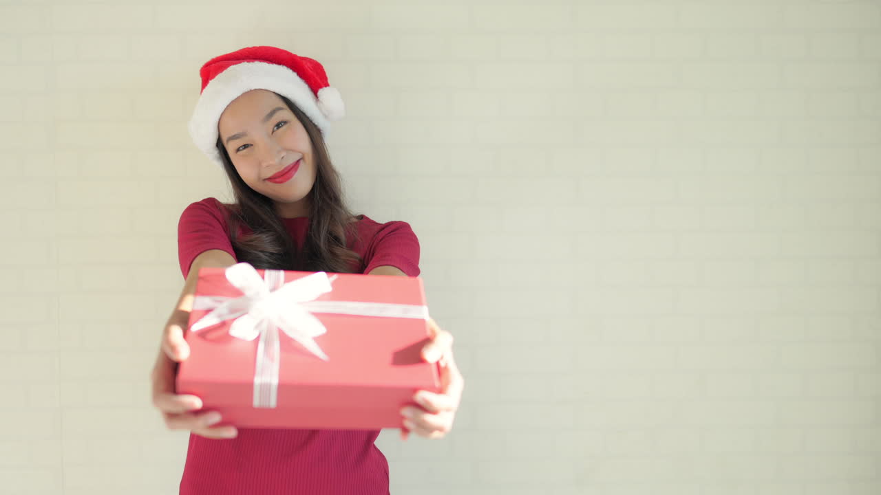 medio ancho de una mujer asiática adulta joven aislada contra un fondo de estudio blanco roto con ropa informal elegante roja y sombrero de navidad sonriendo, ofrece una caja de regalo envuelta en una foto de meme con espacio para copiar