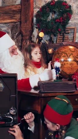 santa claus and children are exploring the country on a retro globe. christmas elves sitting on the floor set up an old gramophone. vertical video