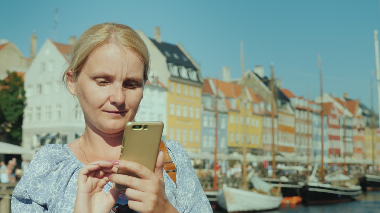 행복한 여성은 스마트폰을 사용하여 운하와 인기 있는 관광 거리의 배경에 서 있습니다.