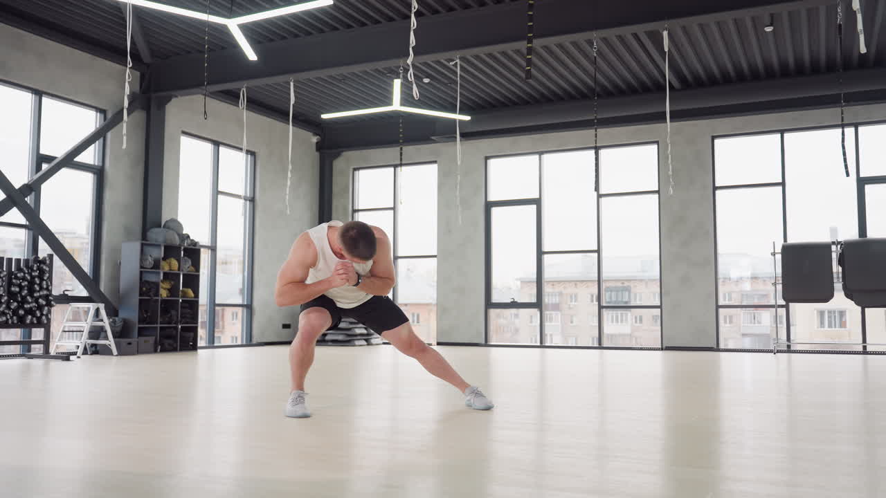 Bodybuilder performing side lunge warm up exercise in bright modern gym with white tank and black shorts balancing on light wooden floor beneath hanging straps and large windows urban