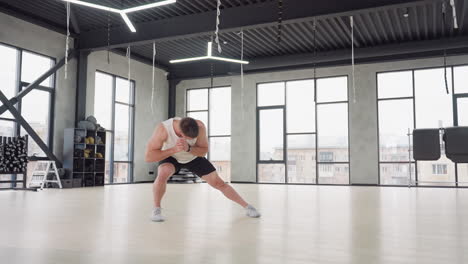 bodybuilder performing side lunge warm up exercise in bright modern gym with white tank and black shorts balancing on light wooden floor beneath hanging straps and large windows urban