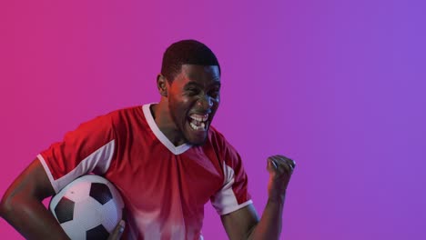 Happy-african-american-male-soccer-player-raising-hand-with-football-over-neon-pink-lighting