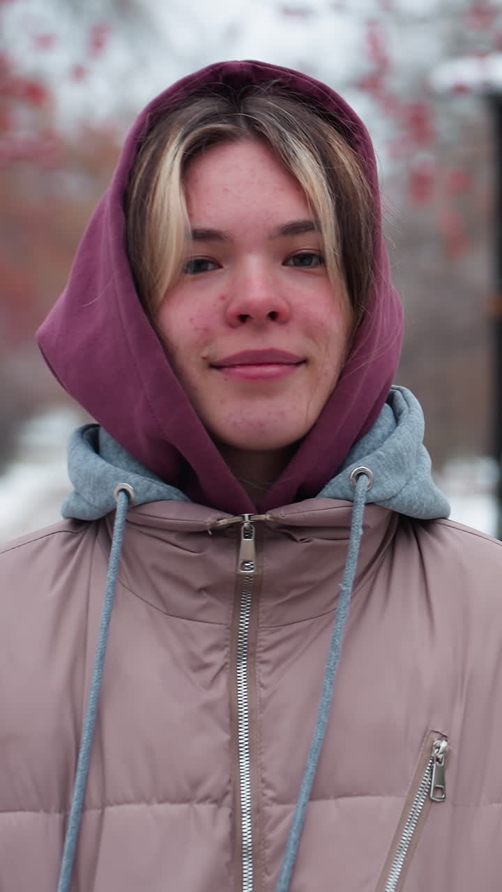 mujer joven con una chaqueta con capucha cálida sonríe con las manos en los bolsillos mientras está de pie en el camino cubierto de nieve, el parque de invierno con árboles de bayas rojas y farolas lejanas