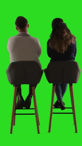 a man and a woman sitting on chairs back to back, with a green screen background