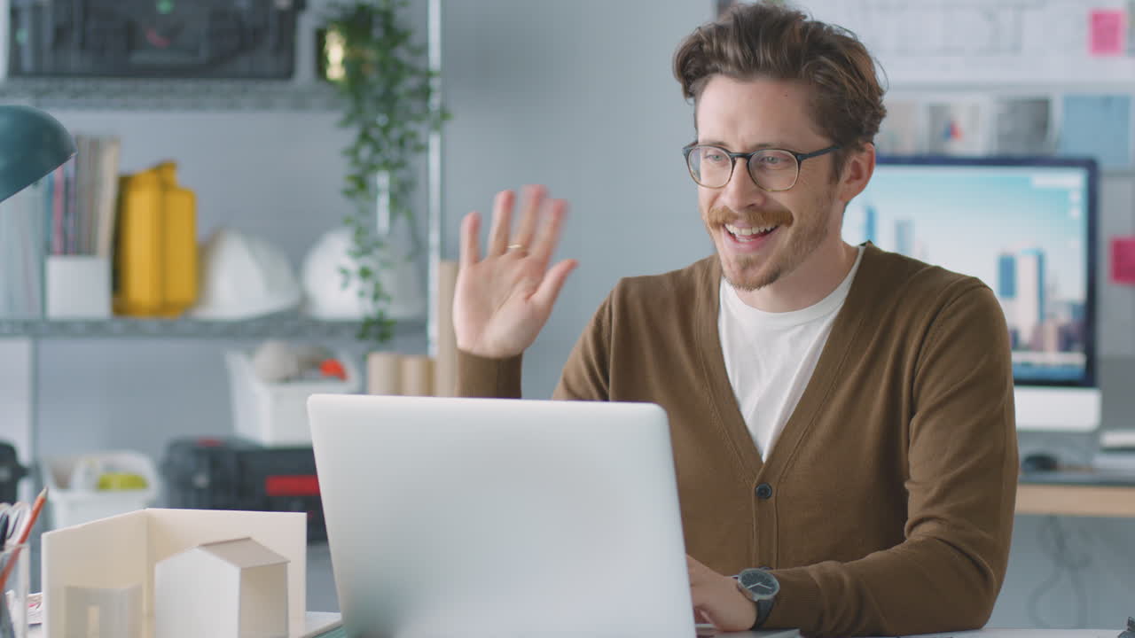 Miesarkkitehti toimistossa pitämässä videopuhelua kannettavalla tietokoneella työpöydän ääressä.