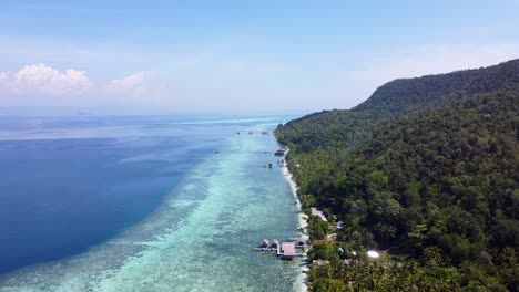 reverse aerial view of beach huts over crystal clear ocean water, coral reefs and rainforest trees on tropical island in raja ampat, west papua, indonesia