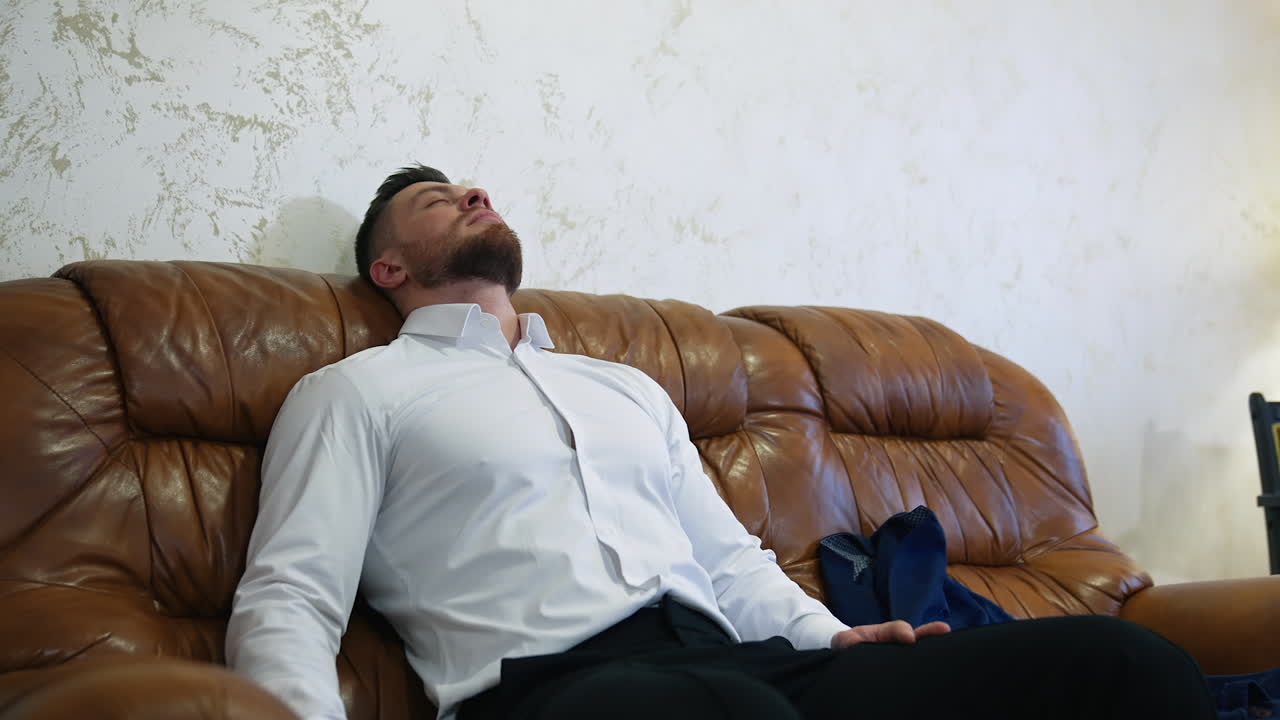 Tired man resting after a long trip. Handsome man in white shirt is sitting on a comfortable couch in a hotel room.