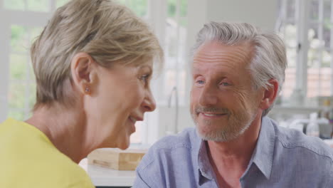 Close-Up-Of-Kissing-Retired-Couple-Sitting-Around-Table-At-Home-Talking-And-Laughing-Together