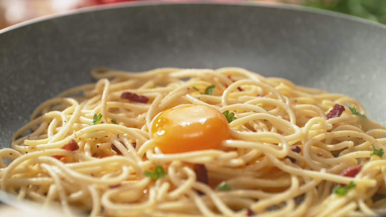 Spaghetti Carbonara in una padella, con un tuorlo d'uovo che cade sui fili caldi della pasta con pancetta ed erbe tritate - Ripresa in allontanamento, al rallentatore.
