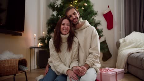 Portrait-of-a-happy-couple-guy-and-girl-in-white-sweaters-sitting-on-the-floor-near-a-pink-gift-hugging-and-looking-at-the-camera-in-a-cozy-Christmas-decorated-house-in-winter