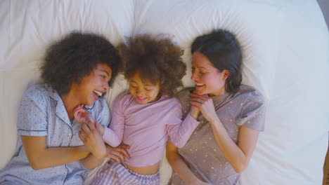 overhead portrait of family with two mums wearing pyjamas playing on bed at home with daughter