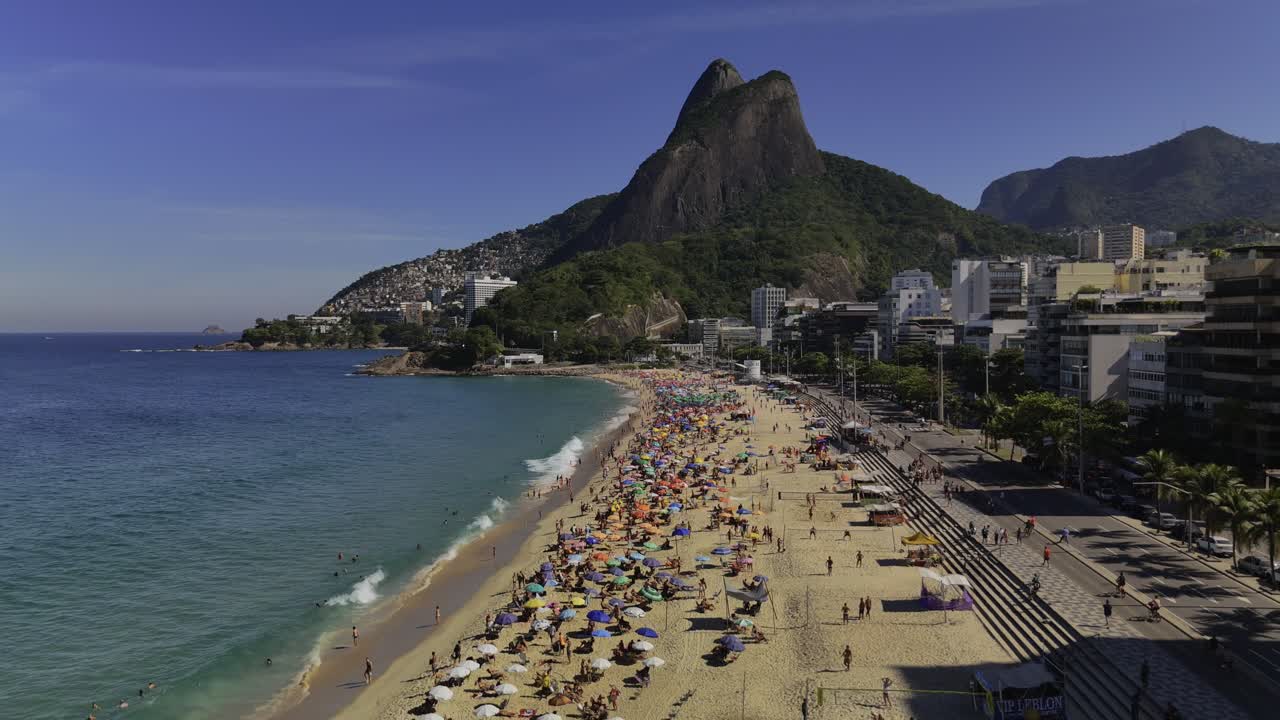 Backward aerial over crowded and bustlin Lebron Beach in Rio de Janeiro, Brazil, overlooking the iconic Pedra do Arpoador