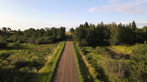 4k drone footage of car driving towards and underneath camera drone on red dirt road between lush green trees during sunset