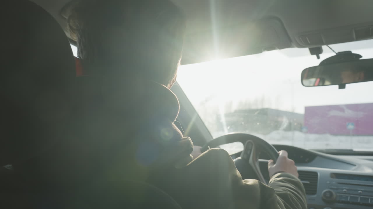 rear view of man dancing in car cabin while steering wheel held, sunlight filtering through windshield onto snow covered parking lot with blurred buildings