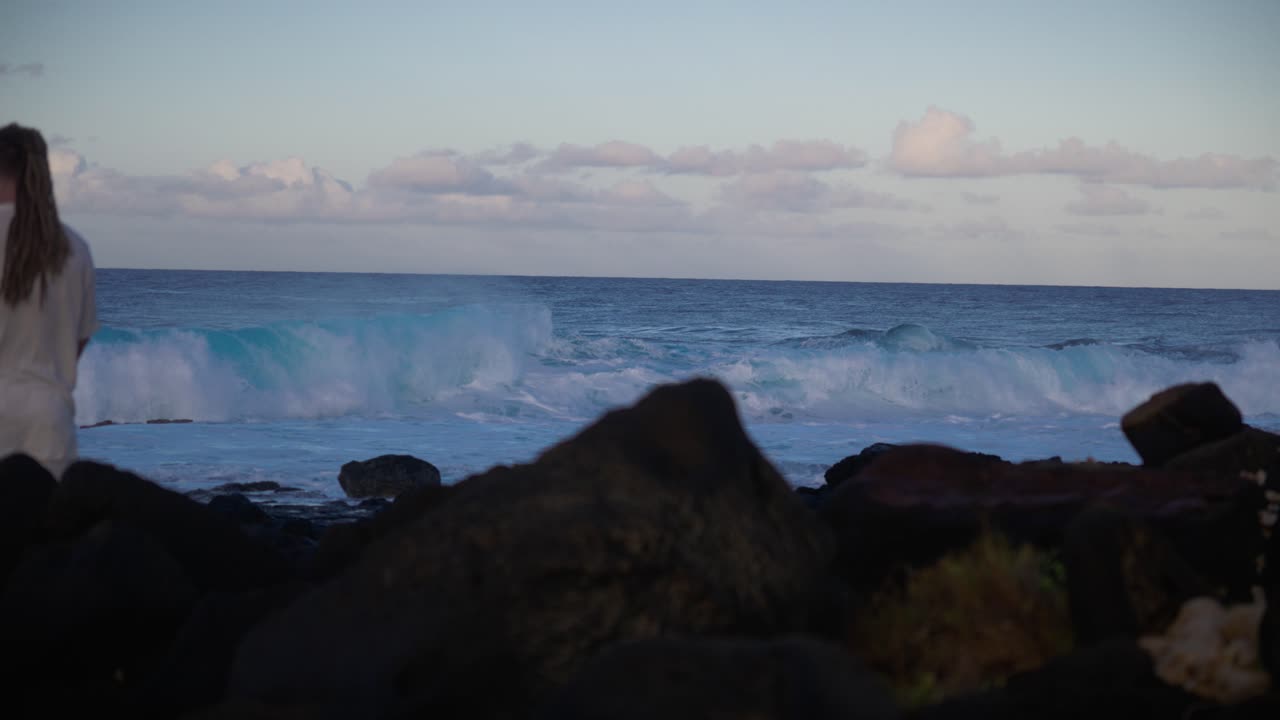 A solitary figure stands quietly on volcanic rocks overlooking the ocean, evoking contemplation and serenity as waves move rhythmically beneath soft evening light