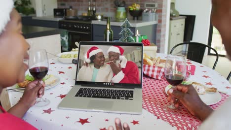 African-american-couple-with-wine-using-laptop-for-christmas-video-call-with-happy-couple-on-screen