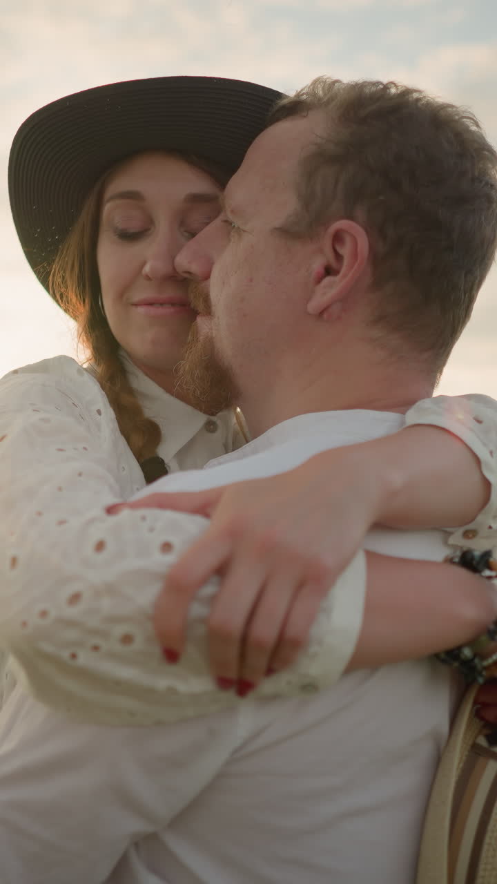 A husband and wife share a tender embrace in a grassy field at sunset, smiling as their faces touch. Both dressed in white outfits, the woman wears a hat and holds another in her hand