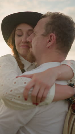 a husband and wife share a tender embrace in a grassy field at sunset, smiling as their faces touch. both dressed in white outfits, the woman wears a hat and holds another in her hand