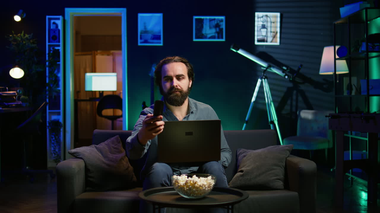 hombre escribiendo en la computadora portátil, sentado en el sofá y viendo la televisión