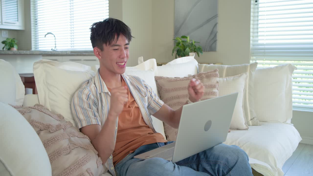 カードを持ったアジア人男性が、ラップトップでカード情報を入力して支払いを行い、ガッツポーズをとりながらソファでくつろいでいる。
