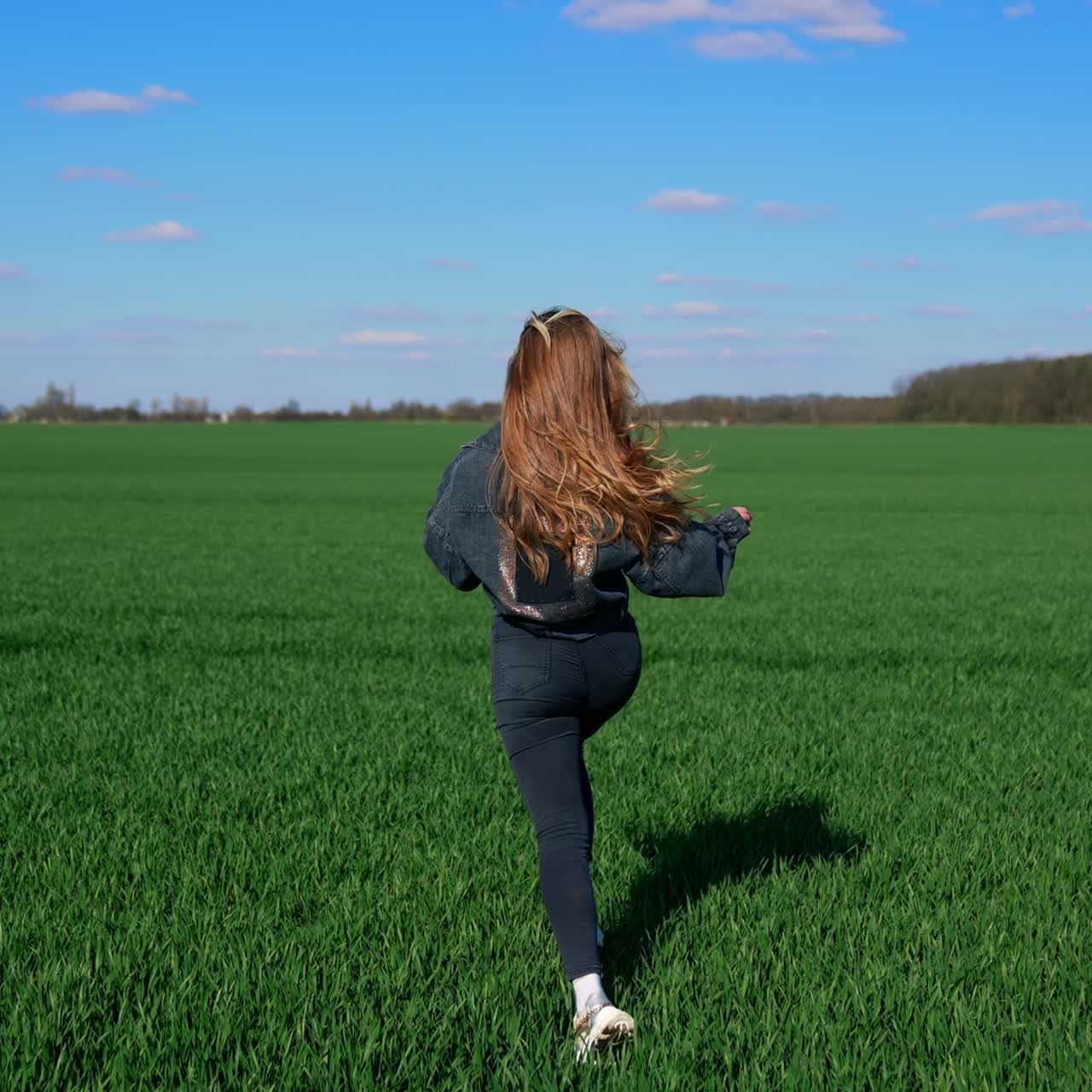Young woman in denim suit runs on field. Happy beautiful girl is running outdoors in bright spring day. Rear view