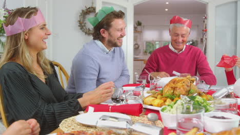familia de varias generaciones con sombreros de papel leyendo chistes de galletas de navidad mientras comen juntos