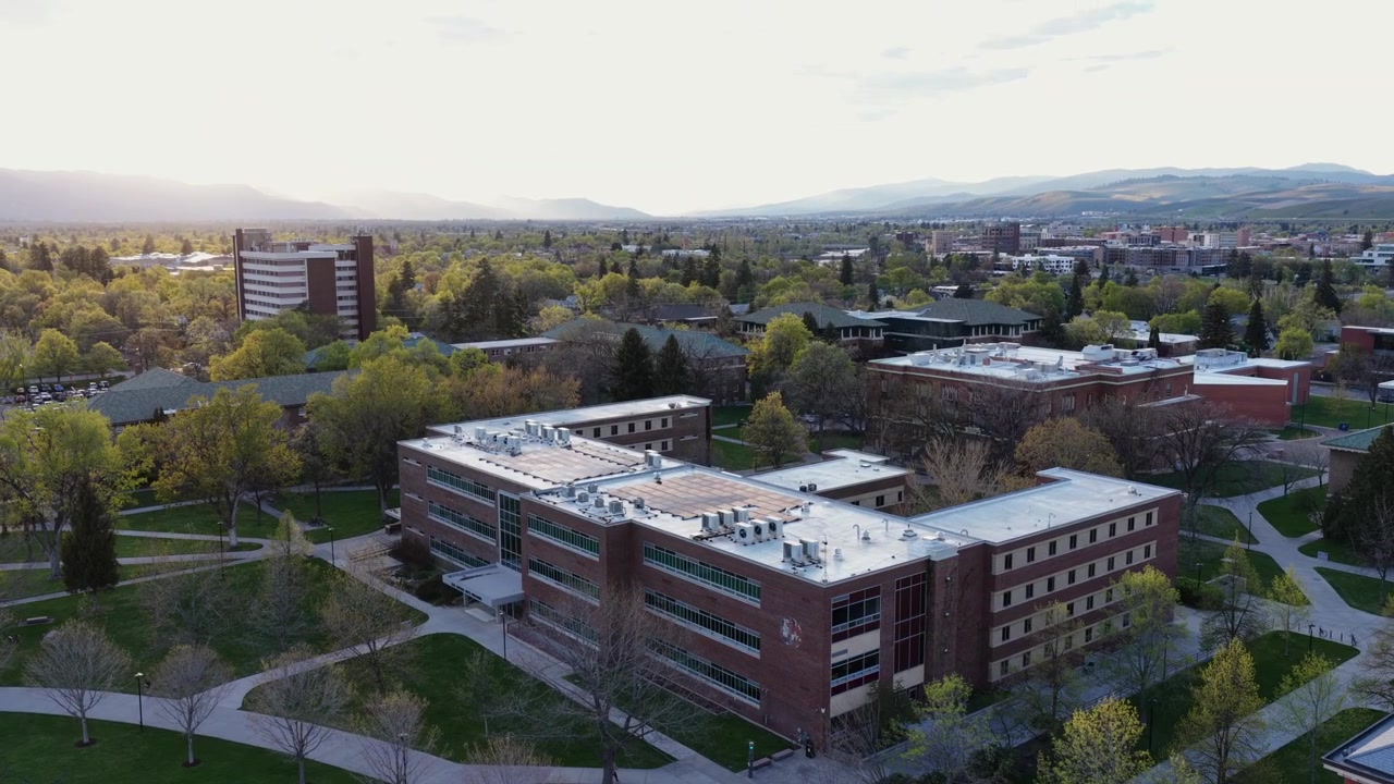 Campus de la Universidad de Montana en Missoula, Montana, EE. UU., con edificios académicos, arquitectura de ladrillo, árboles verdes y senderos en un entorno urbano tranquilo con montañas y luz vespertina. Órbita de dron aérea.