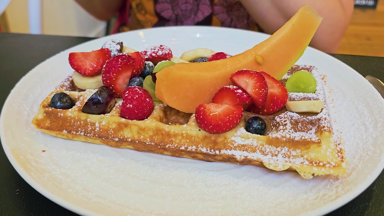 Fresca cialda belga guarnita con fragole, mirtilli, lamponi, banana, uva, melone e zucchero a velo su un piatto bianco in un'accogliente ambientazione da colazione in caffè con calda luce interna.