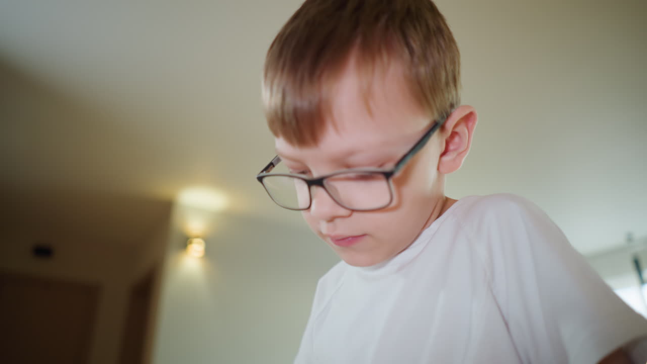 Close up of boy with glasses wearing white t shirt indoors, focused expression while concentrating on activity, natural light illuminating face, background blurred, educational moment at home