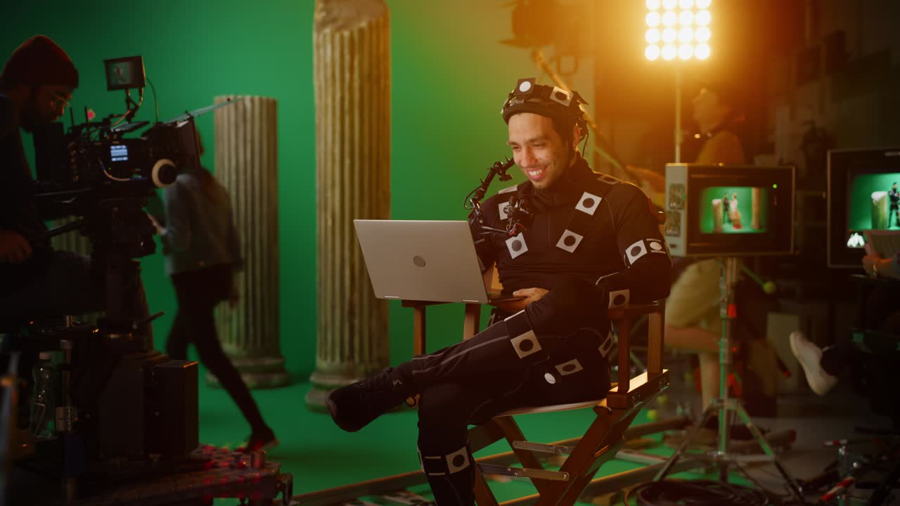 hombre guapo y sonriente con traje de captura de movimiento y equipo de cabeza teniendo descanso para almorzar, sentado en una silla, usando una computadora portátil. estudio película de alto presupuesto. en el estudio de cine escenario de teatro de vestuario