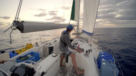 man controlling sails with rope on winch, sailing with sailboat at open sea with sunset on horizon