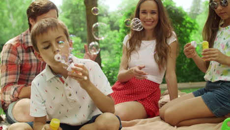 Parents-sitting-on-blanket-with-kids-in-forest.Boy-and-girl-blowing-soap-bubbles