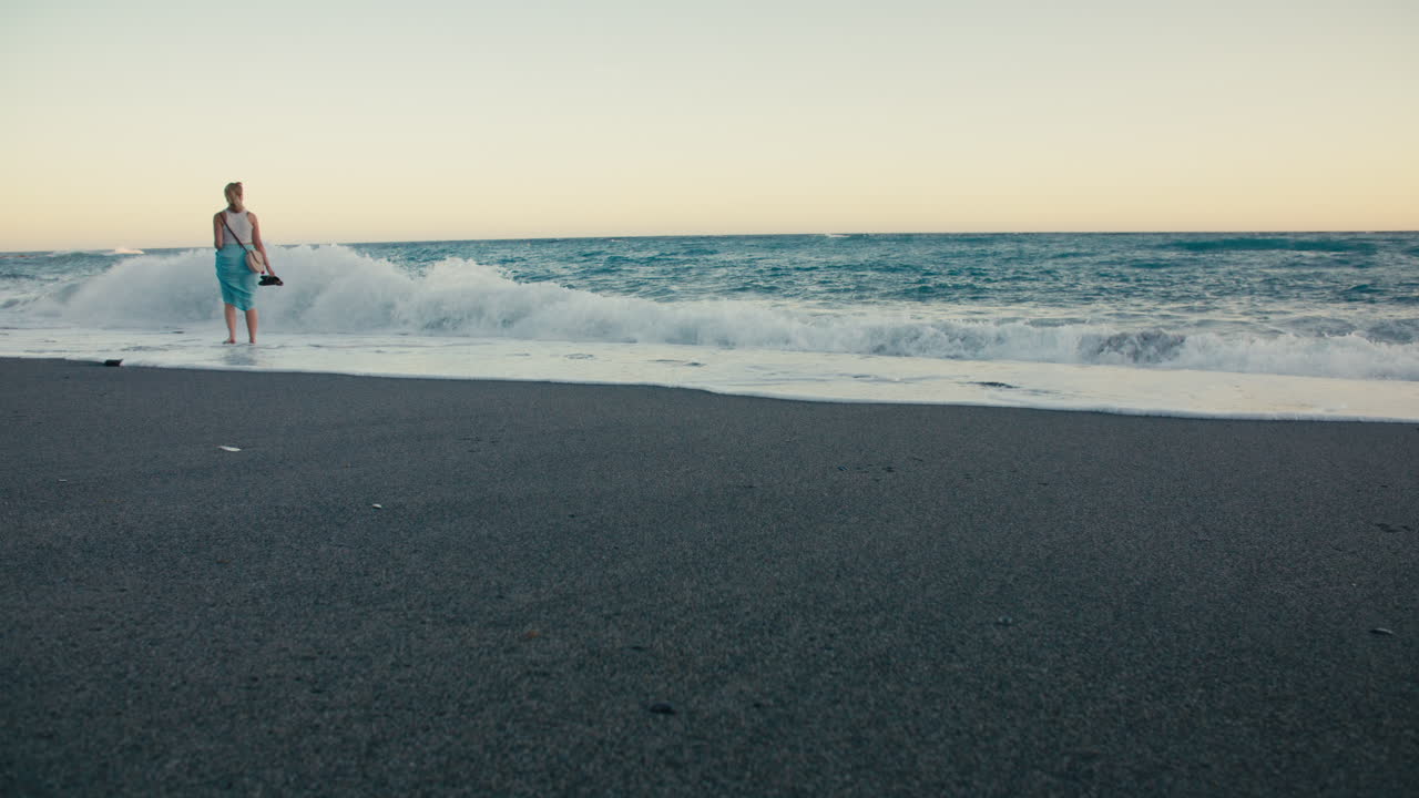 Wide angle shot of a young blonde woman strolls barefoot along the beach, holding her shoes, with soft golden light and waves in the background