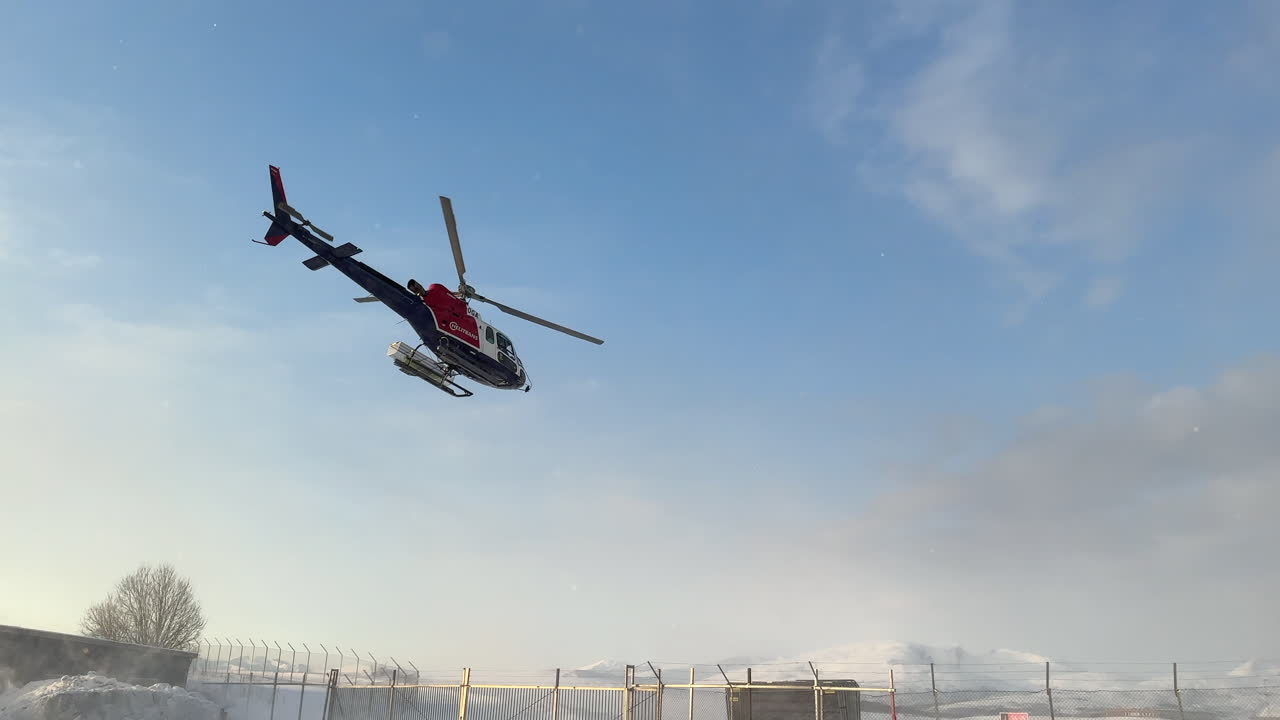 Medium shot of a helicopter on a portable helipad taking off with snow blowing around from the prop wash on clear winters day