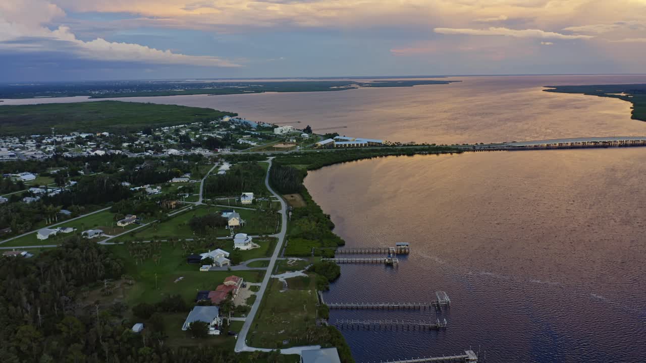 A serene coastal community lines the edge of a golden river, where long wooden piers stretch into the still water beneath a sky painted with soft hues of evening light