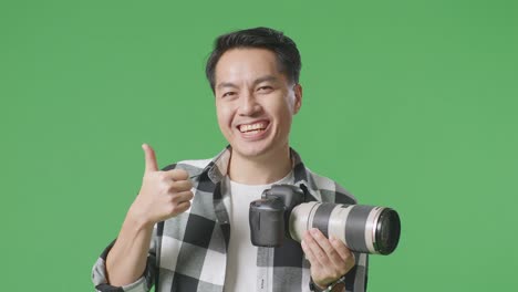 스튜디오에서 녹색 스크린 배경에 서 있는 동안 엄지손가락을 올려놓고 미소 짓는 카메라를 가진 아시아 사진작가의 클로즈업
