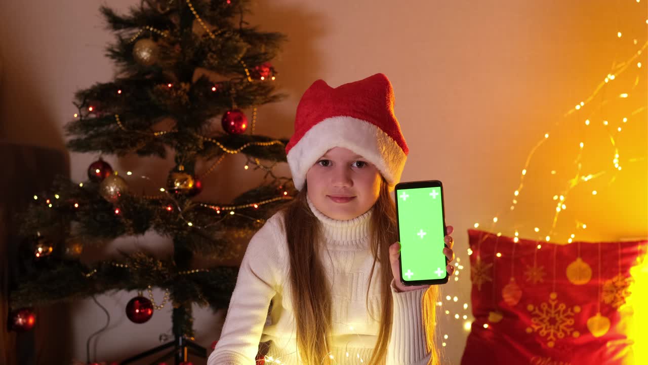 크리스마스 모자를 입은 귀여운 어린 소녀가 녹색 화면으로 휴대전화를 들고 있습니다. 크로마 키 스마트폰은 광고를 위해 설정되어 있습니다. 귀여운 아이는 카메라를보고 있습니다. 디지털 장치를 가진 사랑스러운 아이.