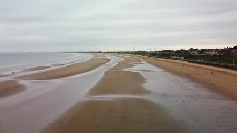 Vista-Aérea-De-Una-Playa-De-Arena-Con-Marea-Baja-Que-Muestra-Turbinas-Eólicas-En-El-Fondo-En-Un-Día-Nublado-Y-Aburrido-En-Bridlington,-Este-De-Yorkshire,-Reino-Unido,-Inglaterra