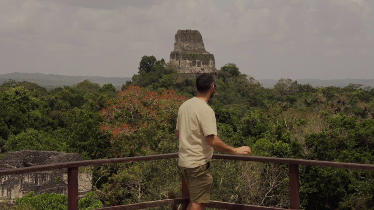 Tourist man in balcony turn to watch Tikal Pyramid temple in Guatemala. Glasses wanderer. Slow motion